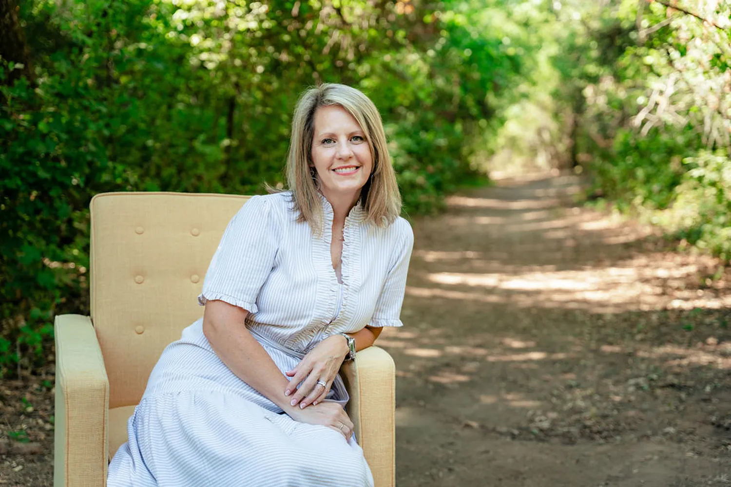 Mamie Bronson seated in a yellow armchair on a wooded path.